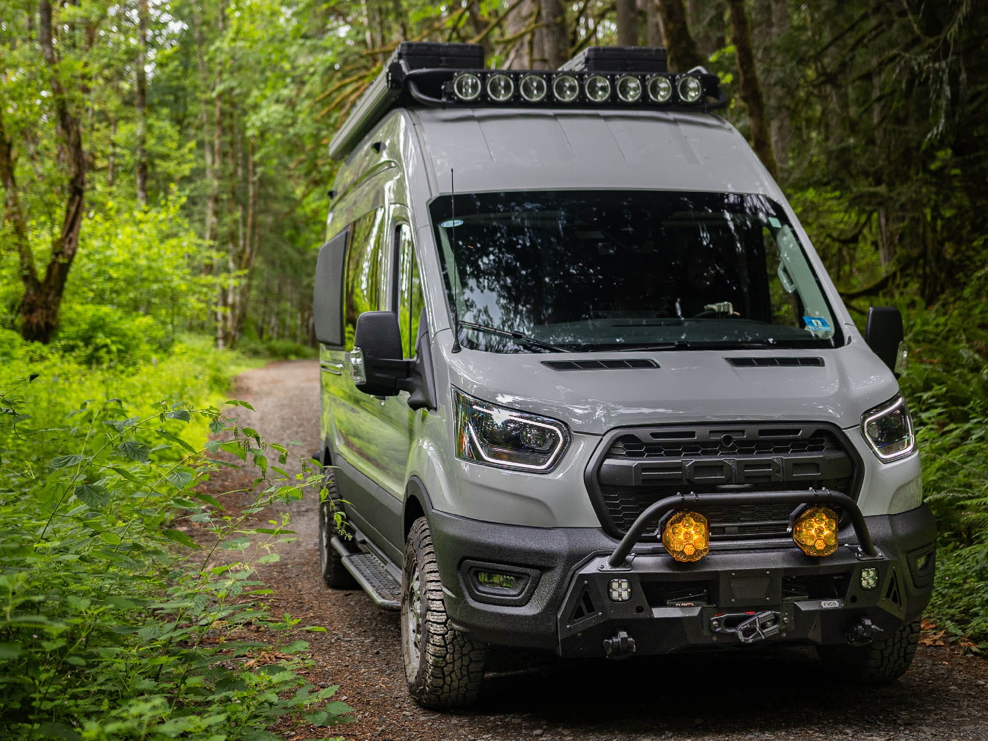 Front view of Ford Transit Van with FVC Transit Side Steps, Safari Roof Rack, and Core Front Bumper installed while driving through the woods