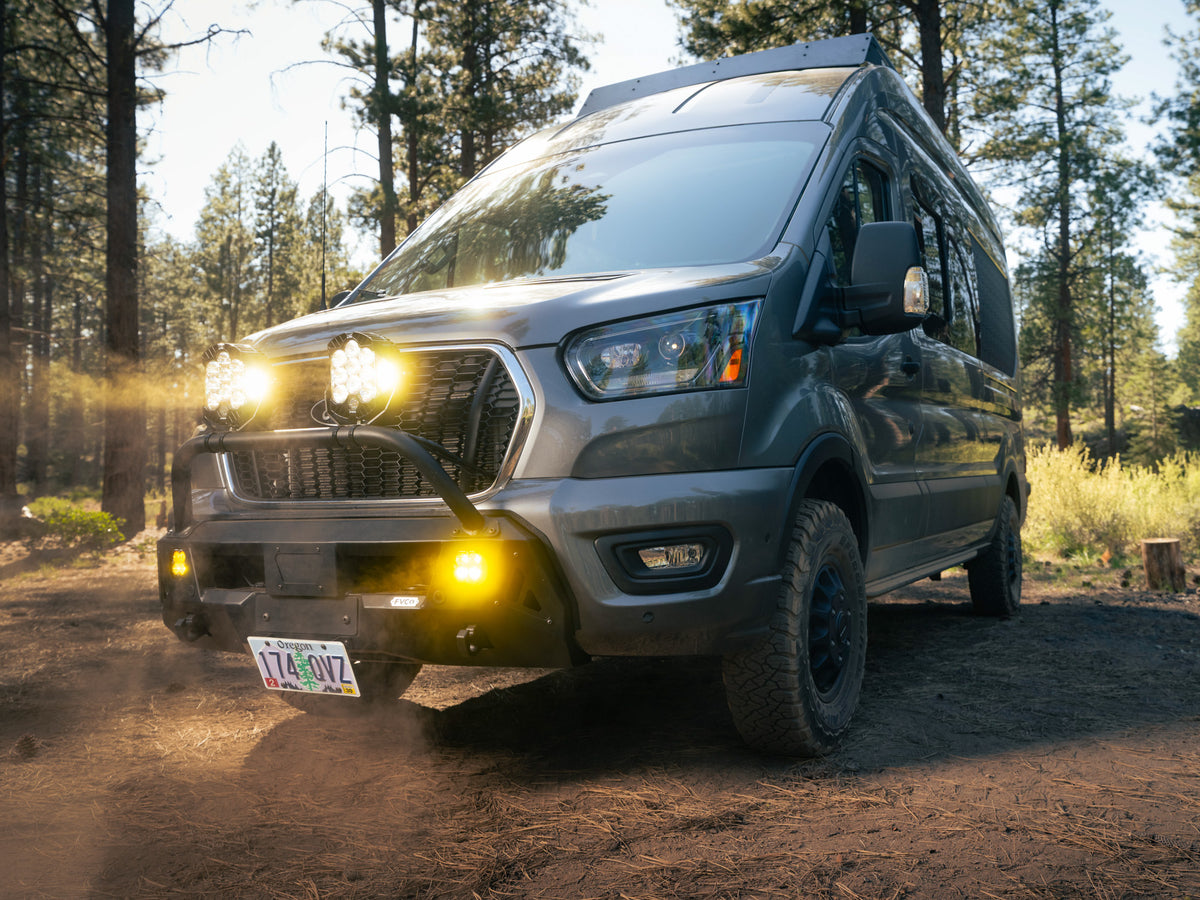 A dark gray off-road Transit van with bright yellow lights and a  FVC Front Bumper with Bull Bar and Skid Plate, parked on a dirt road in a pine forest.