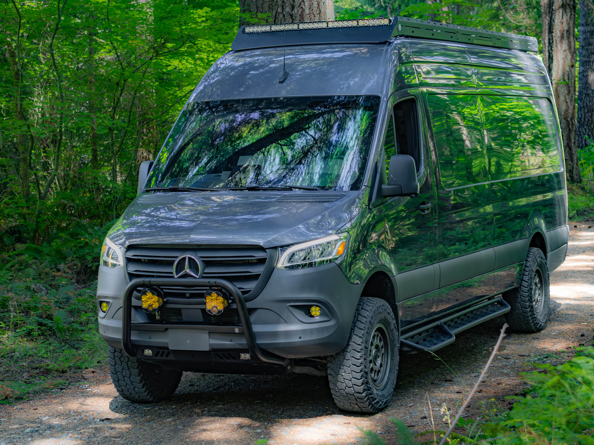 Angled front view of Sprinter adventure van driving through the woods with Sprinter Bull Bar, Skid Plate, Standard Roof Rack, and Side Steps by FVC